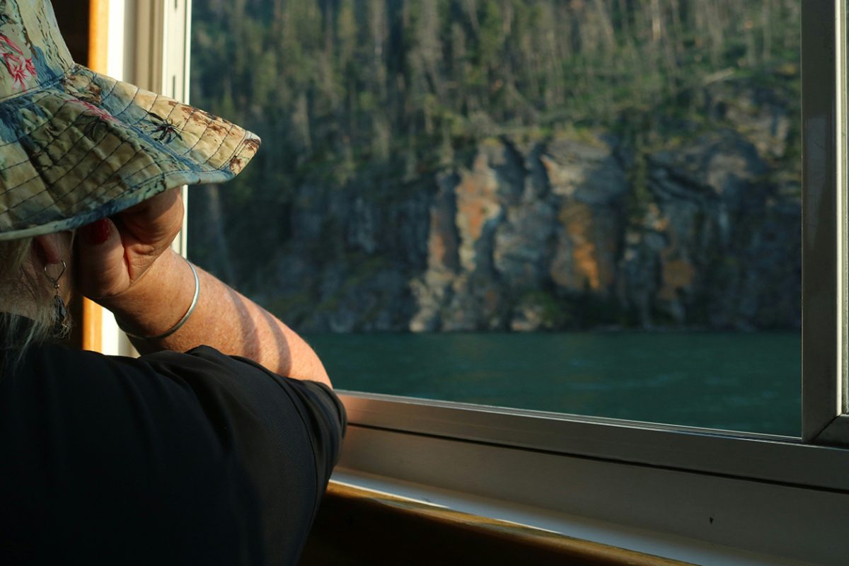 Person in a sun hat looking out a window at a lake and cliffs