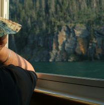 Person in a sun hat looking out a window at a lake and cliffs